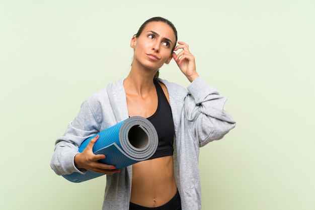 Mujer joven del deporte con la estera sobre la pared verde aislada que tiene dudas y con la expresión de la cara confusa