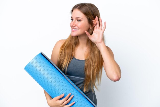 Mujer joven deporte que va a clases de yoga mientras sostiene una estera escuchando algo poniendo la mano en la oreja