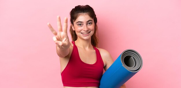 Mujer joven deporte que va a clases de yoga mientras sostiene una estera feliz y cuenta tres con los dedos