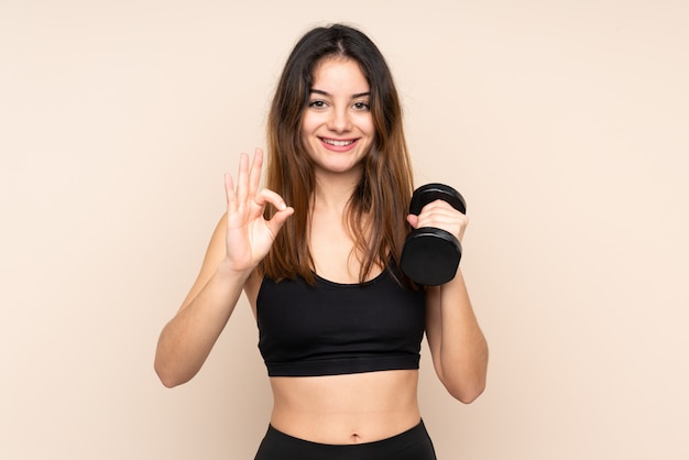 Mujer joven del deporte que hace levantamiento de pesas aislado en la pared beige que muestra una muestra aceptable con los dedos