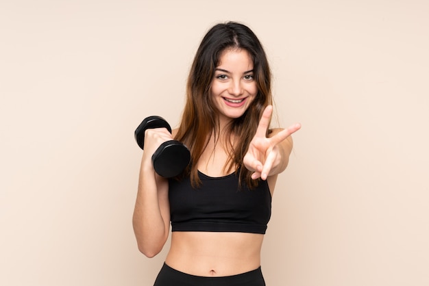 Mujer joven del deporte que hace levantamiento de pesas aislado en la pared beige sonriendo y mostrando el signo de la victoria