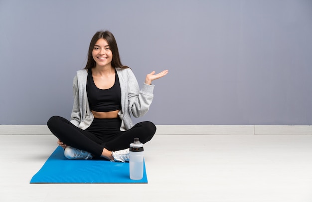 Mujer joven del deporte que se sienta en el piso con la estera que sostiene el copyspace imaginario en la palma