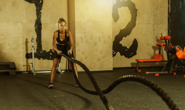 Mujer joven deporte en ropa deportiva está entrenando con cuerda de batalla en el gimnasio. Entrenamiento funcional. Concepto de estilo de vida saludable