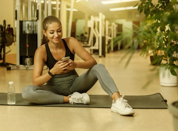 Mujer joven deporte en ropa deportiva con smartphone mientras está sentado en la colchoneta en el gimnasio. Pausa de entrenamiento. Concepto de estilo de vida saludable