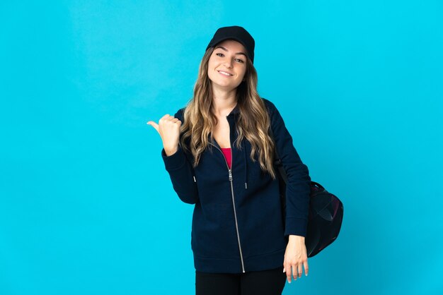 Mujer joven deporte rumano con bolsa de deporte aislado en la pared azul apuntando hacia el lado para presentar un producto