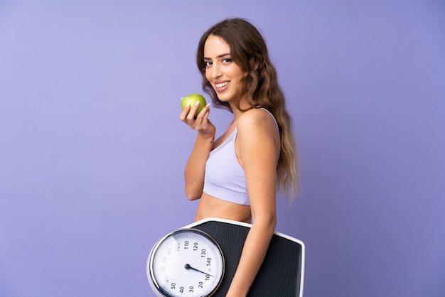 Mujer joven del deporte sobre la pared púrpura aislada con la máquina de pesaje y con una manzana