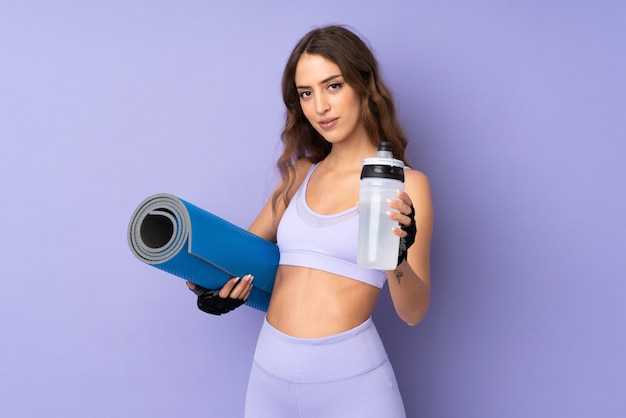 Mujer joven deporte sobre pared púrpura con botella de agua deportiva y con una estera