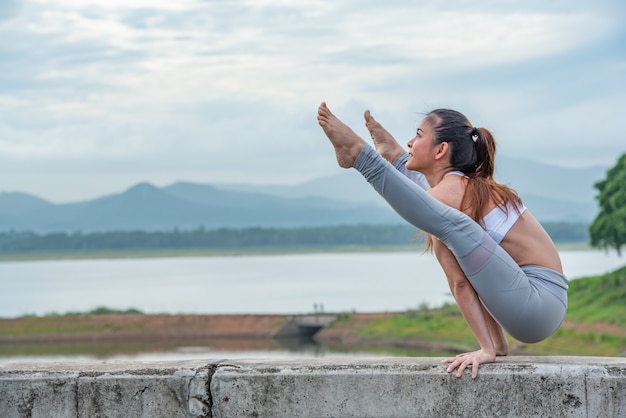 Mujer joven de los deportes que hace yoga en el lago con el fondo de la montaña.