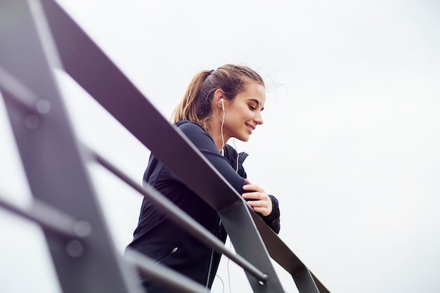 Mujer joven deportiva con los auriculares que se inclinan en la cerca y que miran lejos al aire libre