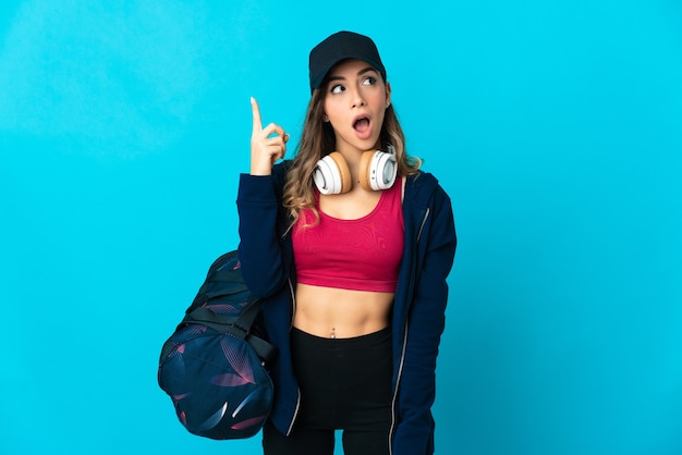 Mujer joven deportiva con bolsa de deporte aislada en la pared azul con la intención de darse cuenta de la solución mientras levanta un dedo