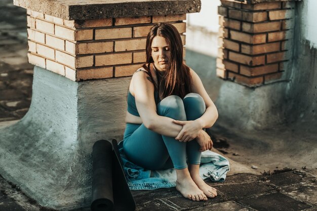 Mujer joven en depresión sentada con las rodillas abrazadas perdidas en malos pensamientos pesimistas y preparándose para entrenar en una terraza en la azotea.