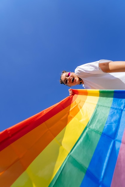 Una mujer joven desarrolla una bandera del arco iris contra el cielo