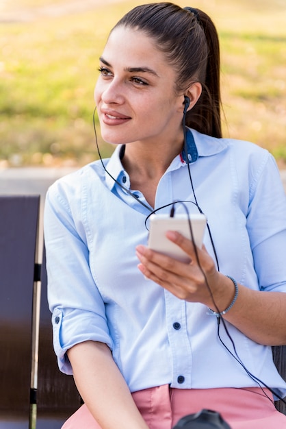 La mujer joven descansa en el parque del otoño en el banco después de un paseo y escucha la música en el teléfono.