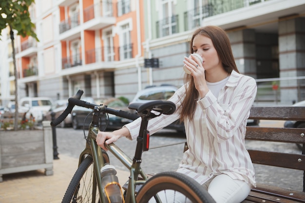 Mujer joven descansando después de andar en bicicleta en la ciudad, tomando café, espacio de copia
