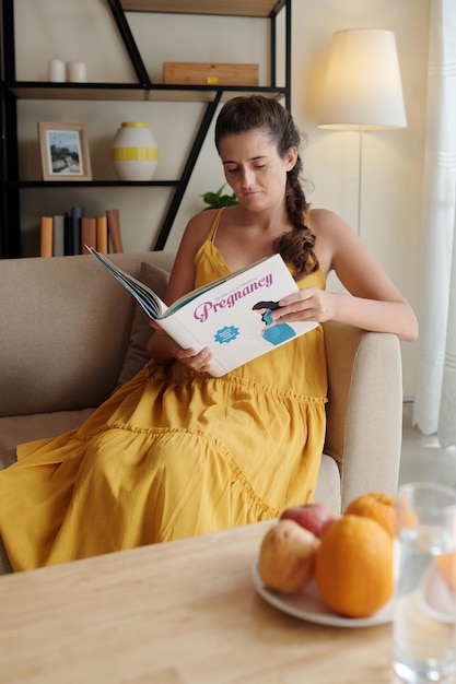 Mujer joven descansando en el sofá en casa y leyendo un libro sobre el embarazo