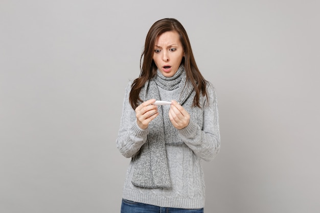 Mujer joven desconcertada en suéter gris, bufanda sosteniendo, mirando el termómetro aislado sobre fondo de pared gris. Estilo de vida saludable, tratamiento de enfermedades enfermas, concepto de estación fría. Simulacros de espacio de copia.