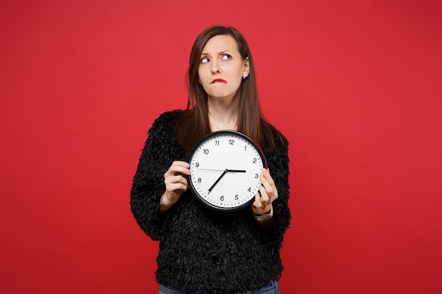 Foto mujer joven desconcertada en suéter de piel negra mirando hacia arriba, mordiendo los labios, sosteniendo el reloj redondo aislado sobre fondo rojo brillante de la pared en el estudio. personas sinceras emociones, concepto de estilo de vida. se acaba el tiempo.