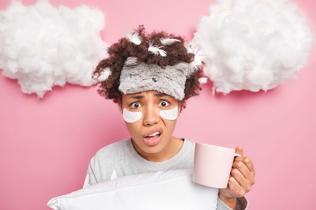 Mujer joven descontenta y disgustada tiene plumas en el cabello desordenado disfruta de una taza de café fresca sostiene una almohada aislada sobre una pared rosa