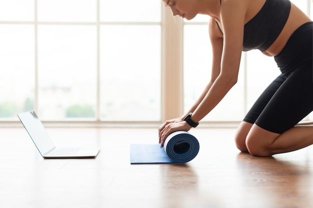 Mujer joven desenrollando estera de yoga en el piso cerca de la computadora portátil