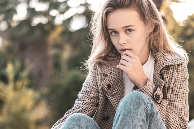 Mujer joven despreocupada en moda vintage pantalones y abrigo sentado en el parque. Linda chica rubia de buen humor posando en día de otoño, disfrutando de buen tiempo.