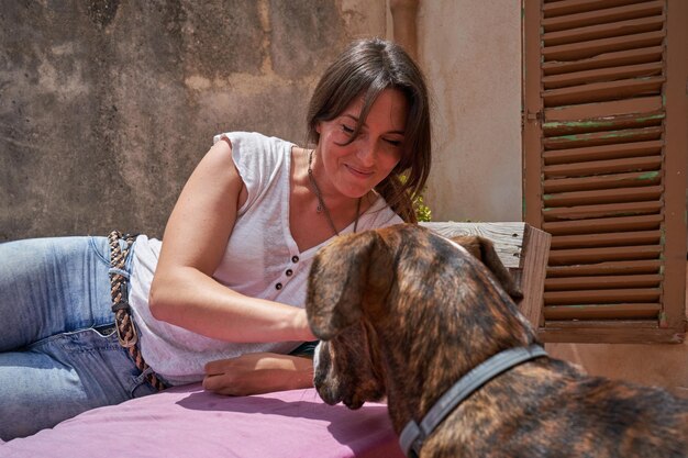 Mujer joven despreocupada con ropa informal descansando en un banco de madera con colchón morado mientras acaricia a un perro de pura raza en la terraza de una casa de piedra el día de verano