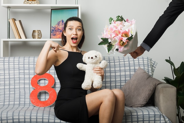 Mujer joven en el día de la mujer feliz sentado en el sofá sosteniendo y apunta al oso de peluche toma ramo por chico en la sala de estar