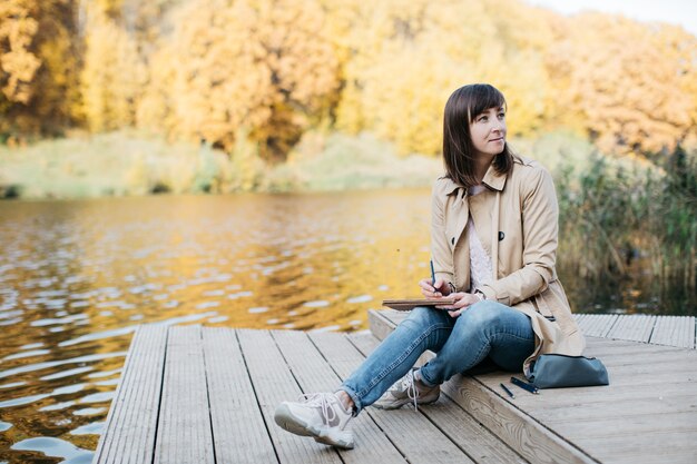 Mujer joven dibujando cerca de un lago en el bosque de otoño