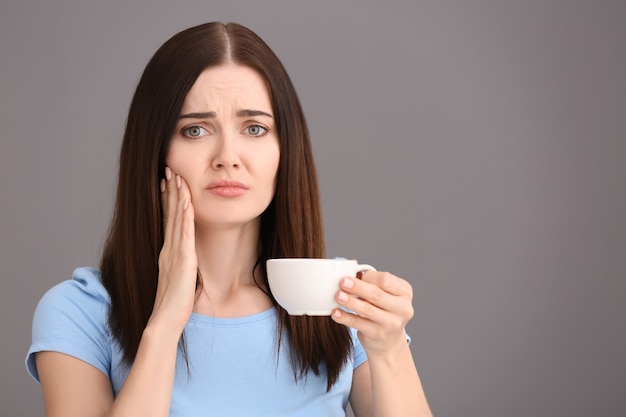 Mujer joven con dientes sensibles y taza de café caliente en gris