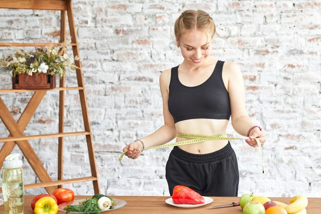 Mujer joven a dieta, mide su cintura con una cinta métrica suave, verduras frescas en la mesa