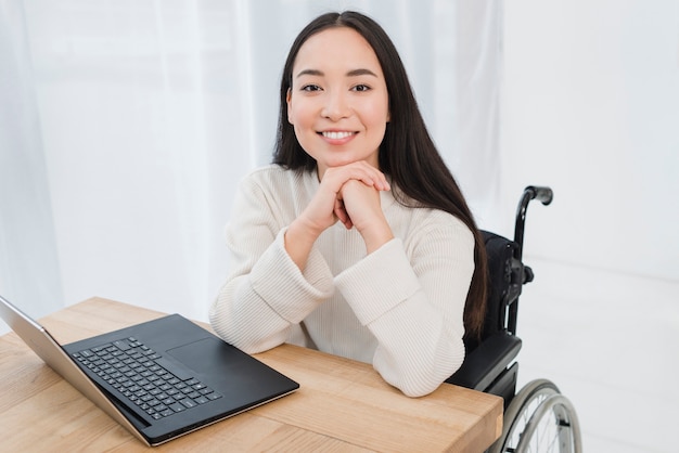 Foto mujer joven discapacitada alegre que se sienta en la silla de ruedas que mira la cámara con el ordenador portátil en la tabla de madera