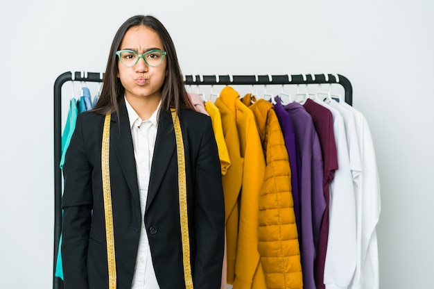 La mujer joven del diseñador asiático aislada en el fondo blanco sopla las mejillas, tiene expresión cansada. Concepto de expresión facial.