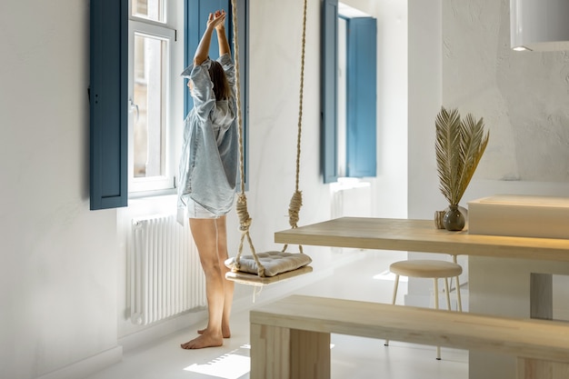 Mujer joven disfruta de la mañana, de pie junto a la ventana en una luminosa sala de estar en un apartamento moderno. Buenos días y concepto de hogar cómodo.