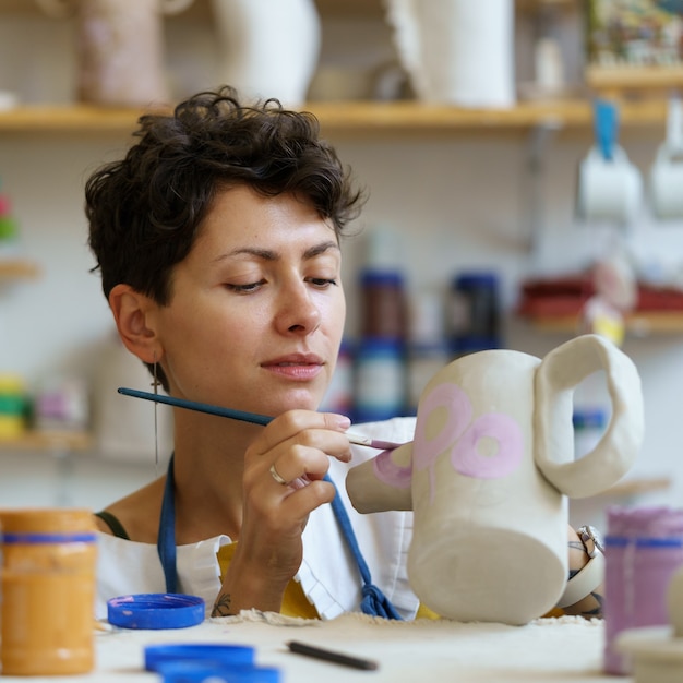Mujer joven disfruta pintando jarra de arcilla con pintura después de dar forma y esculpir la vasija de alfarero de barro