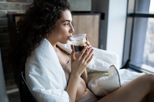 Mujer joven disfrutando de una taza de café sentado junto a una ventana en una mañana soleada