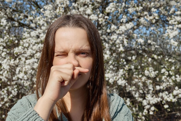 Foto mujer joven disgustada con síntomas de alergia por el polen de los árboles que florecen en primavera, con secreción nasal y estornudos, frotándose la nariz y haciendo caras por sentimientos desagradables.