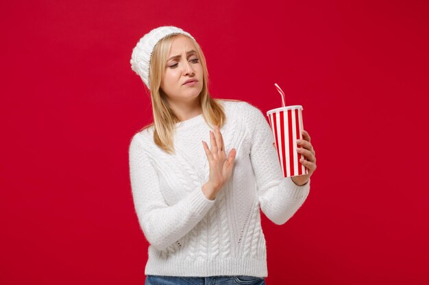 Mujer joven disgustada con suéter blanco, sombrero aislado en el fondo de la pared roja. Estilo de vida de moda saludable, concepto de temporada fría. Simulacros de espacio de copia. Mostrando gesto de parada con la palma de la mano a una taza de cola o refresco.