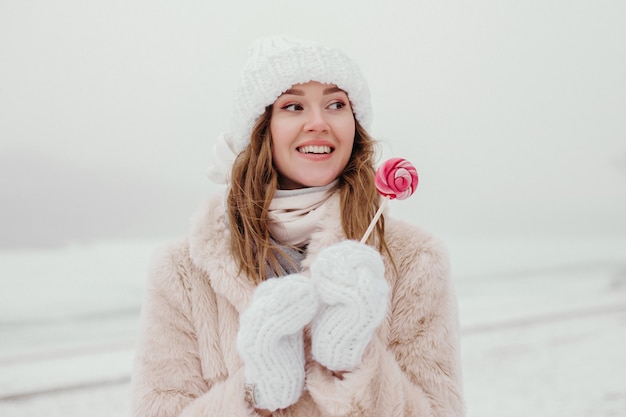 Mujer joven divertida con un abrigo de piel, sombrero blanco y guantes sostiene una paleta en sus manos y sonríe y mira hacia otro lado en el fondo de un bosque de invierno