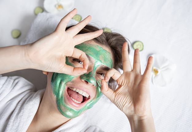 Mujer joven divertida con una máscara cosmética verde en su rostro está descansando mientras está acostado en el salón de spa, vista superior.