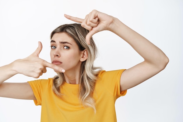 Mujer joven divertida mirando a través de marcos de mano con expresión facial insegura buscando el ángulo perfecto para la foto midiendo algo de pie sobre fondo blanco