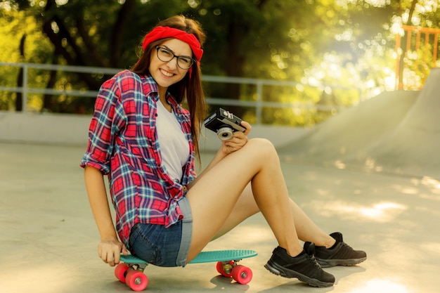 Mujer joven divertida con ropa elegante se sienta en una patineta y utiliza una cámara retro en el skatepark en un día soleado. Concepto de juventud