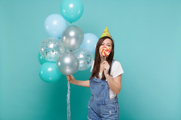 Mujer joven divertida con sombrero de cumpleaños que cubre la boca con piruleta redonda celebrando sostener globos de aire coloridos aislados en el fondo de la pared azul turquesa. Fiesta de cumpleaños, concepto de emociones de la gente.