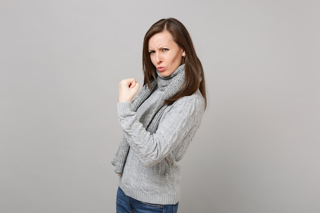 Mujer joven divertida en suéter gris, bufanda mostrando bíceps, músculos aislados sobre fondo de pared gris en estudio. Gente de estilo de vida de moda saludable emociones sinceras, concepto de estación fría. Simulacros de espacio de copia.