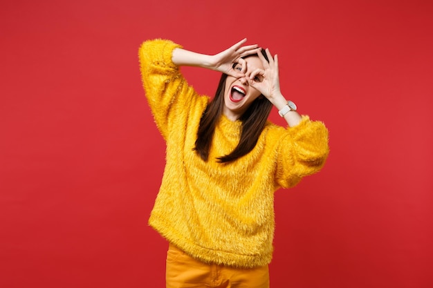 Mujer joven divertida en suéter de piel amarillo cogidos de la mano cerca de los ojos, imitando anteojos o binoculares aislados sobre fondo rojo brillante de la pared. Personas sinceras emociones, concepto de estilo de vida. Simulacros de espacio de copia.