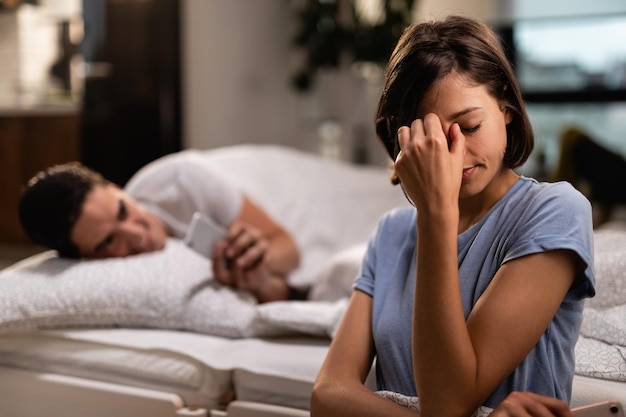 Mujer joven con dolor de cabeza sosteniendo su cabeza con dolor mientras su novio infiel está enviando mensajes de texto en el teléfono móvil a sus espaldas