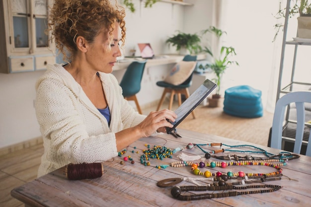 Una mujer joven de edad madura en casa haciendo joyas y collares para la actividad comercial en línea Mujeres sentadas en la mesa en el apartamento haciendo abalorios actividad de ocio interior sola con tableta