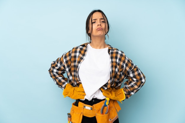 Mujer joven electricista en pared azul enojado