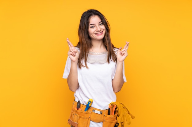 Mujer joven electricista sobre pared amarilla con cruzar los dedos