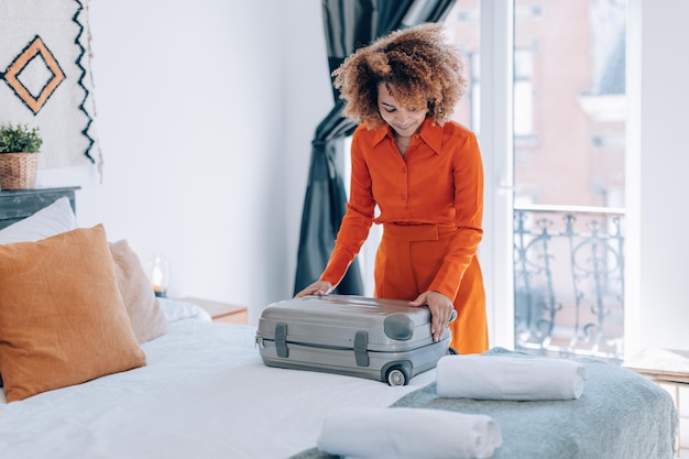 Mujer joven elegante brasileña de pelo rizado en traje naranja abre su equipaje en la cama de la habitación del hotel