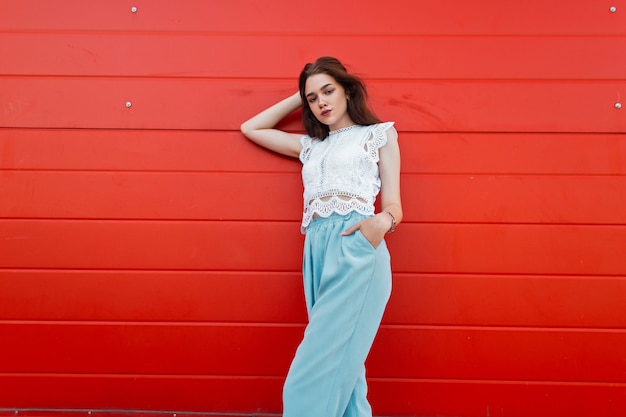 Mujer joven elegante europea en un top de encaje de moda en pantalones  elegantes azules está de pie cerca de un edificio rojo de madera