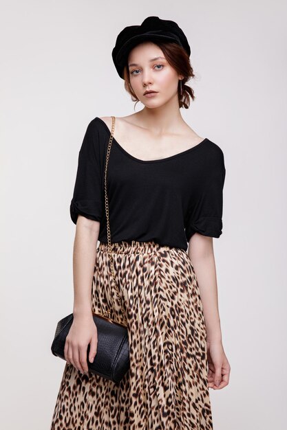 mujer joven elegante en falda larga de leopardo, top negro, sombrero, bolso de mano sobre fondo blanco